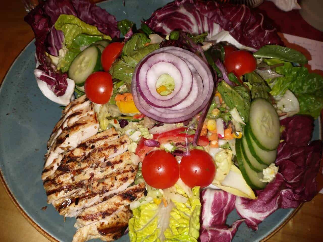 Harzer Schnitzelkönig - Fresh Royal-Salat mit gegrillter Hähnchenbrust, appetitlich angerichtet auf großen, roten Salatblättern und verziert mit Gurkenscheiben, Tomatenstücken und Zwiebelringen